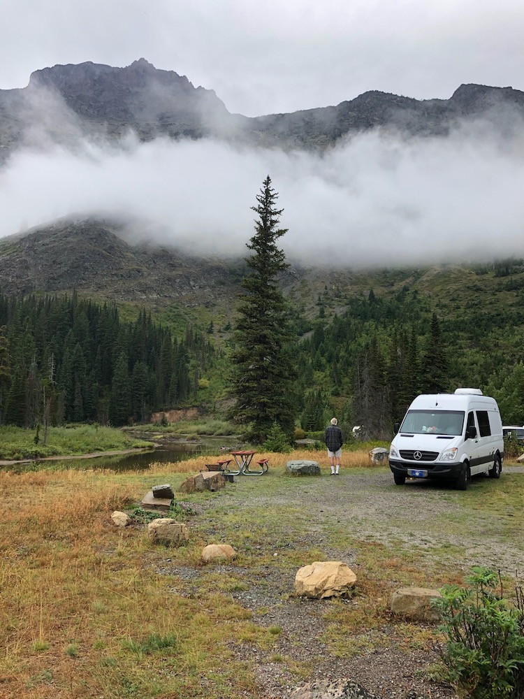 White Mercedes Sprinter converted van parked in the woods, mountains in the background.