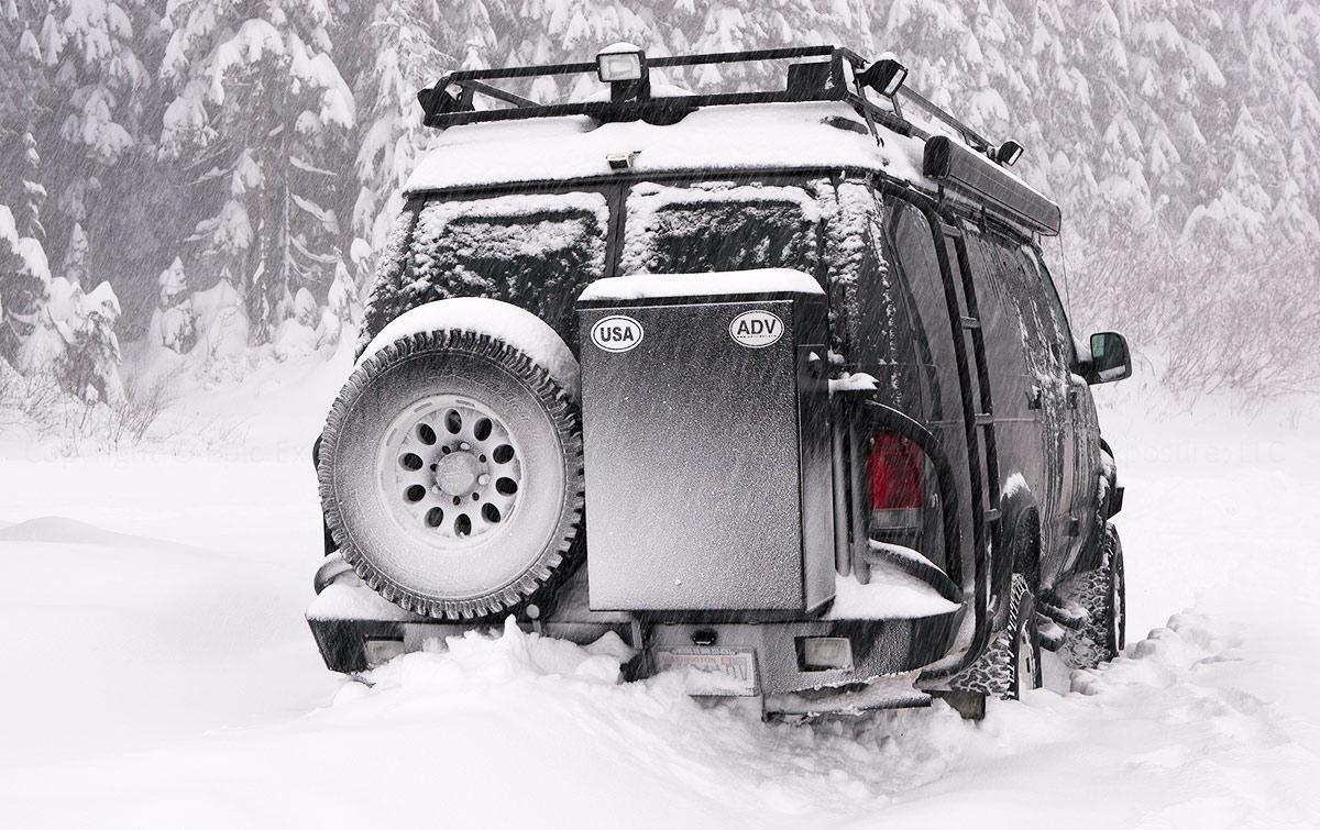 smb-winter Black converted van driving through a forest on a snowy day.
