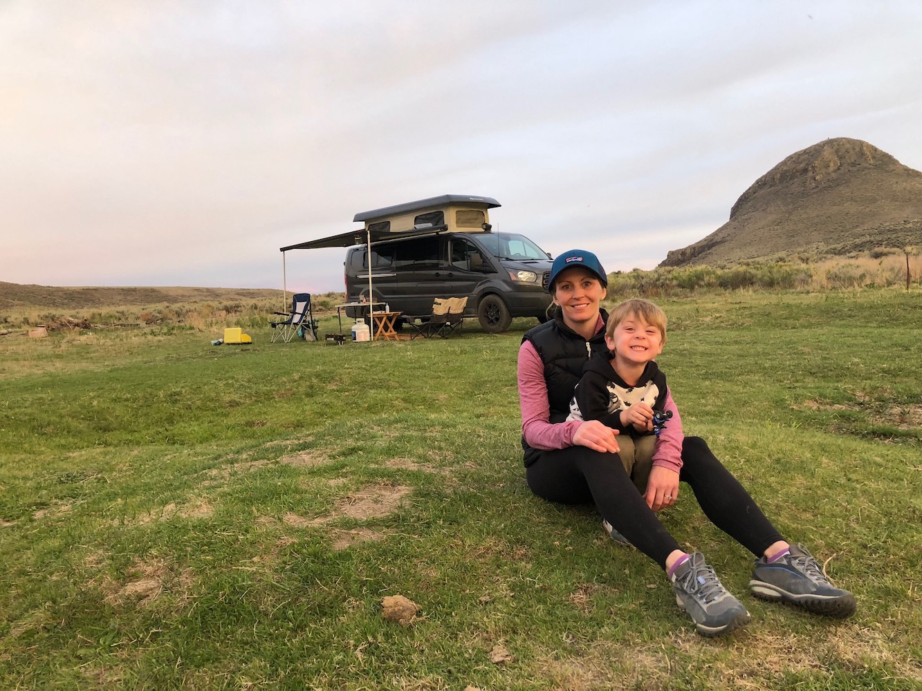 Mother and her son take a picture in the field. Their black converted van parked in the field in the background.