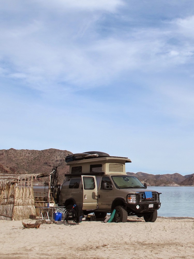 Grey converted van with a penthouse top parked on the beach.